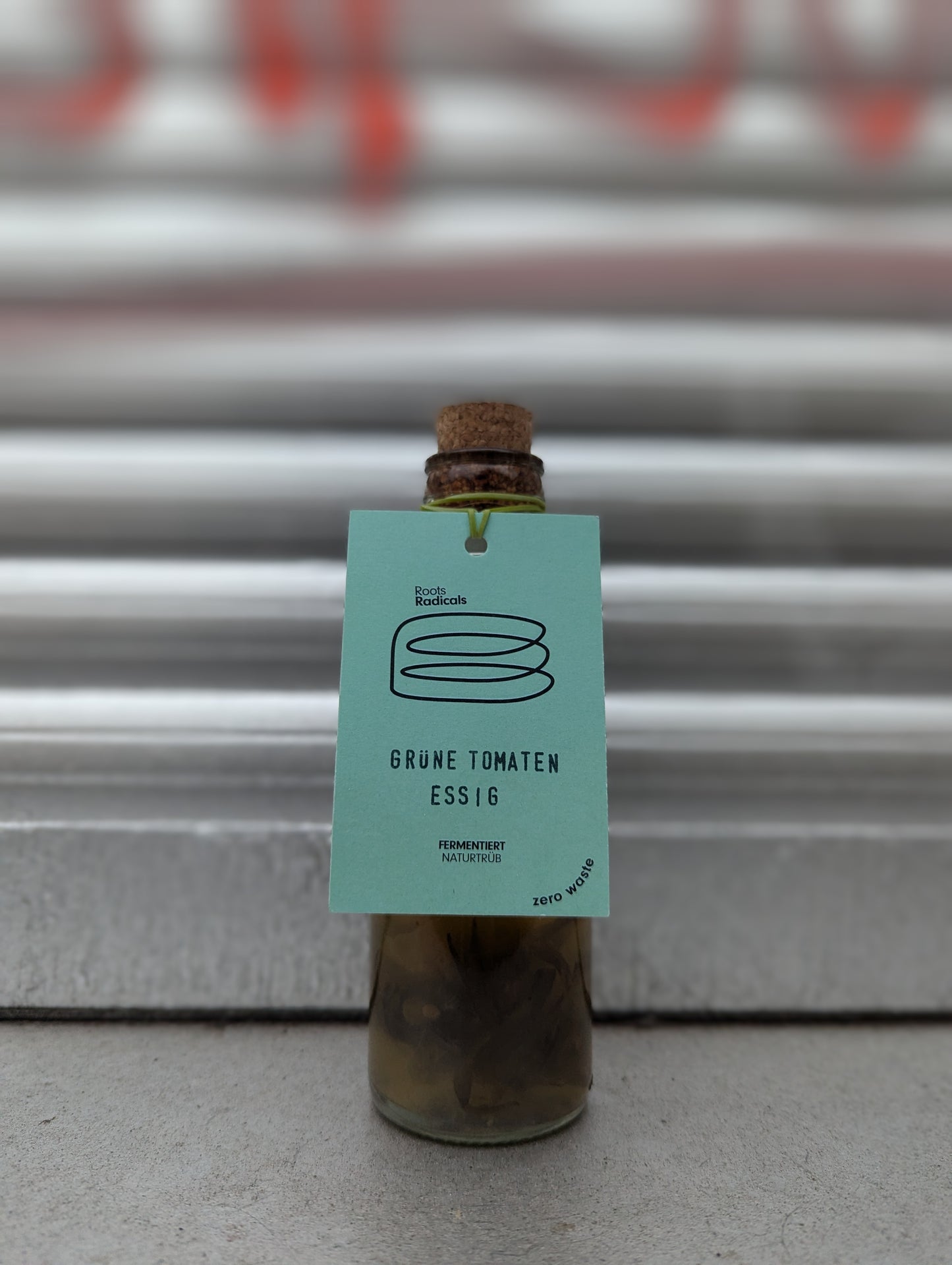 Small glass bottle with a cork stopper, containing a dark liquid and solid pieces. The bottle has a light green tag labeled "Roots Radicals" with a minimalist jar illustration. The text on the tag reads "GRÜNE TOMATEN ESSIG," "FERMENTIERT NATURTRUB," and "zero waste." The bottle is placed against a metallic, industrial background with hints of red graffiti.