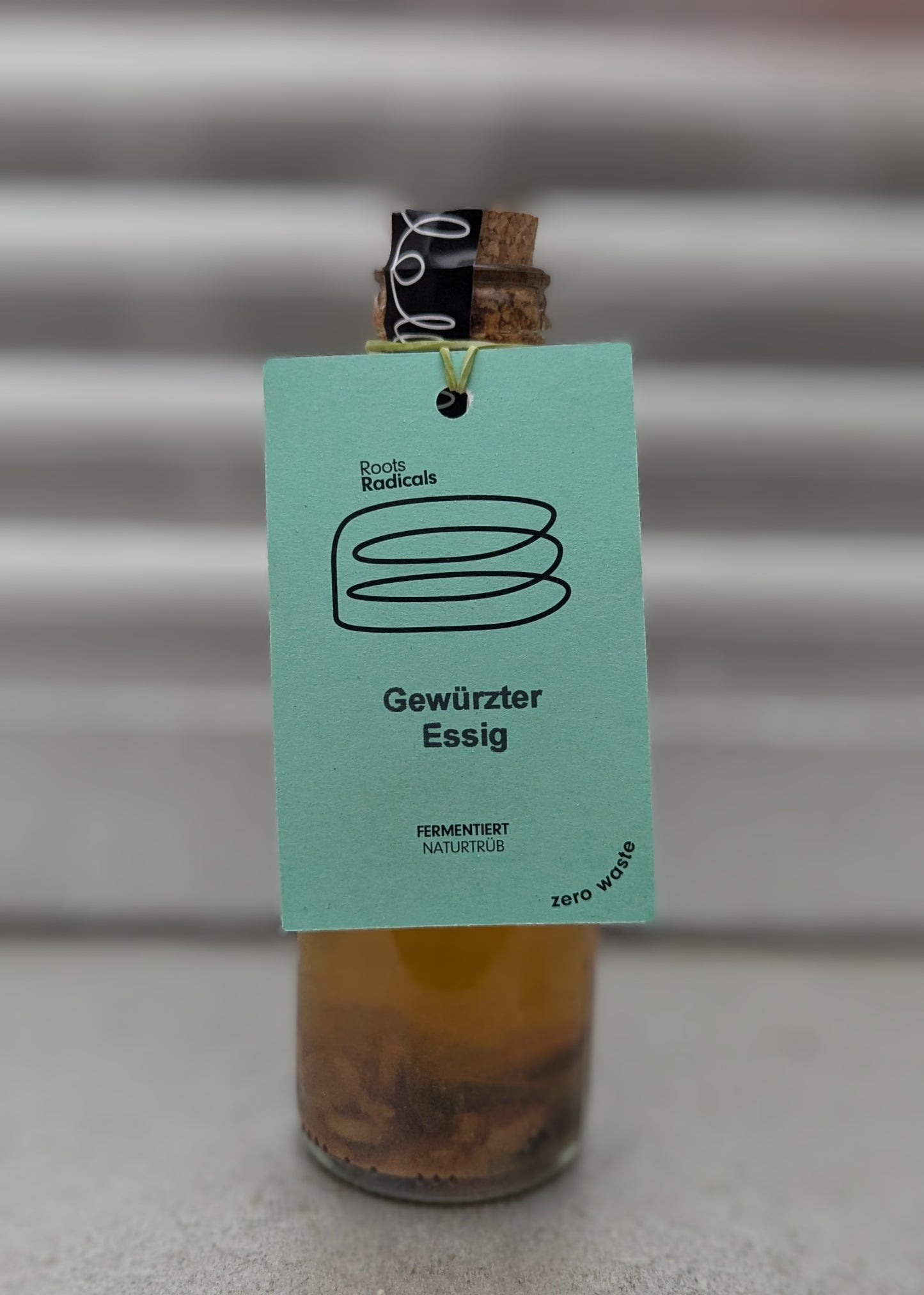 Small glass bottle with a cork stopper, containing a yellowish liquid with solid pieces. The bottle has a light green tag labeled "Roots Radicals" with a minimalist jar illustration. The text on the tag reads "Gewürzter Essig" "FERMENTIERT NATURTRÜB," and "zero waste." The background is metallic with a soft gradient of gray tones.