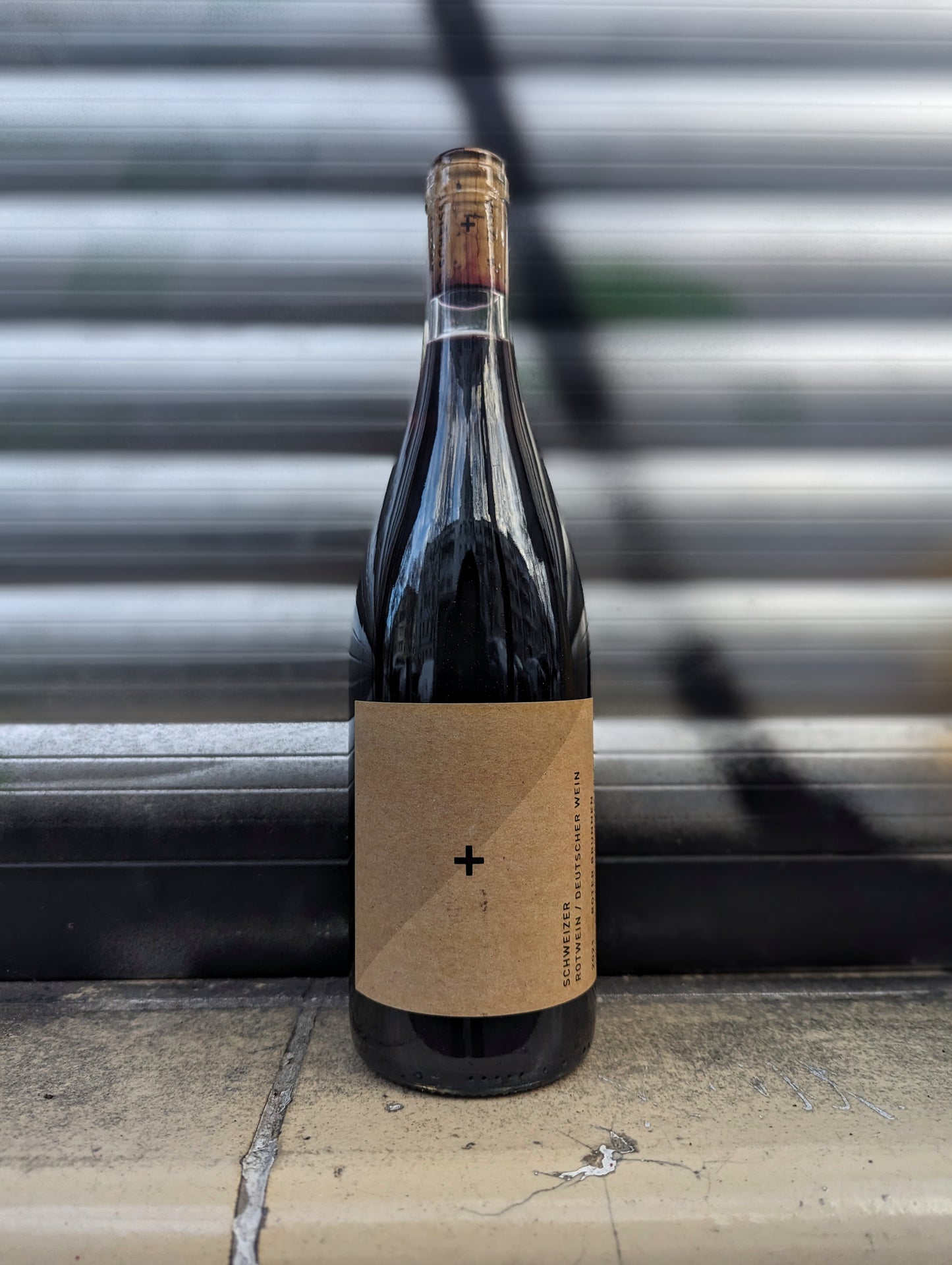 Red wine bottle with minimalist kraft paper label set against a metallic shutter with graffiti accents in an urban Berlin setting.