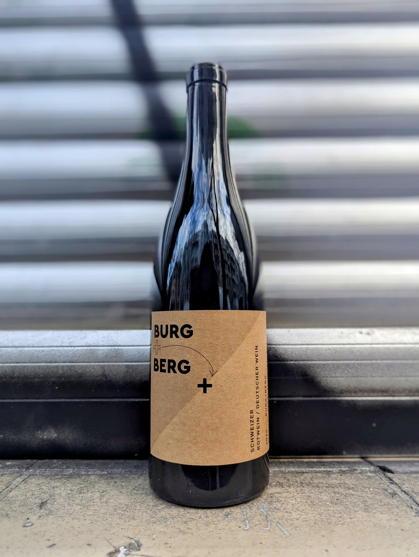 Minimalist wine bottle labeled 'Burg + Berg' with a kraft paper design, set against a metallic shutter background in an urban Berlin setting.