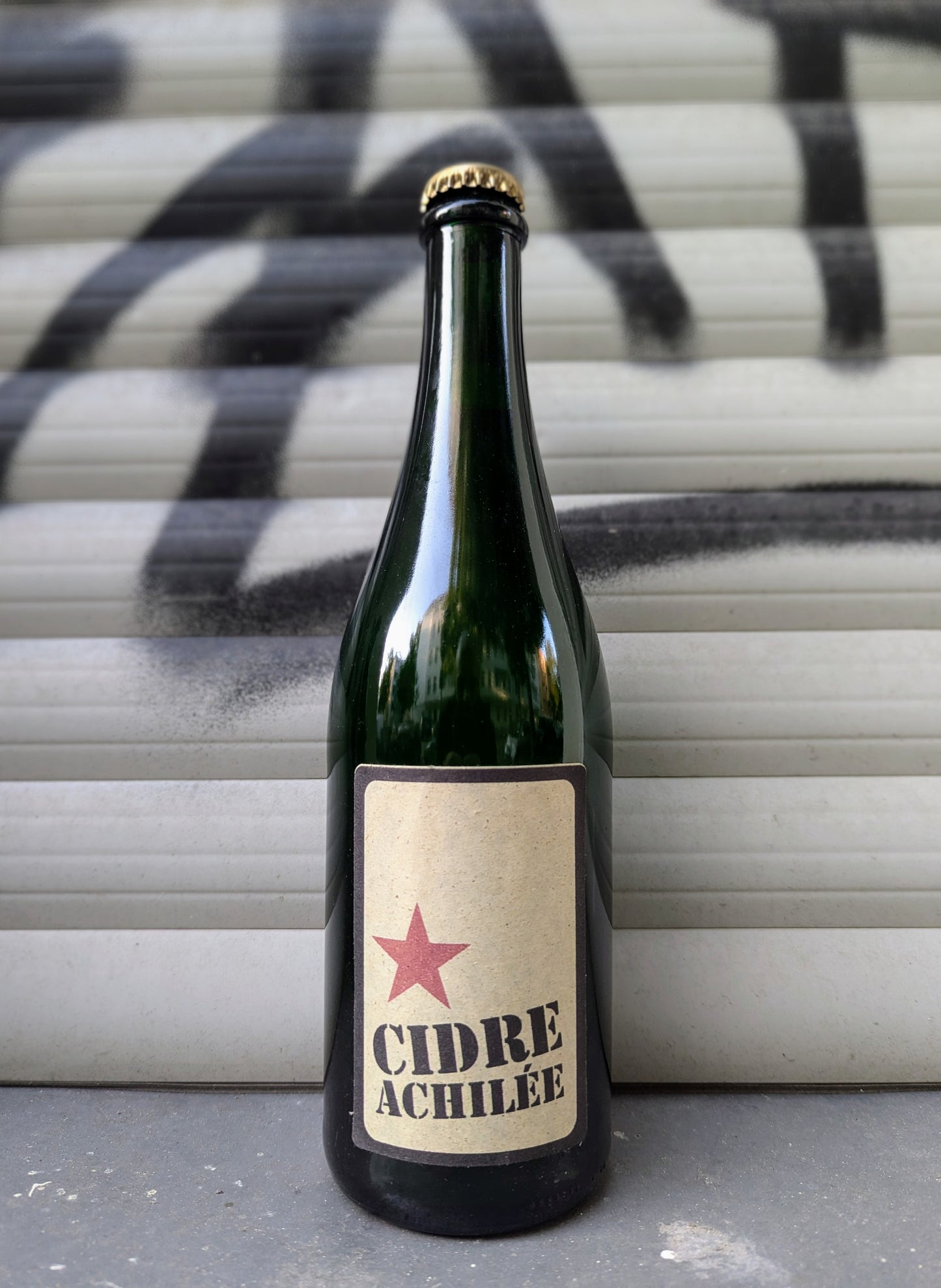 Cidre Achilée cider bottle with a minimalist kraft paper label with a red star set against a graffiti-covered shutter in an urban Berlin setting.