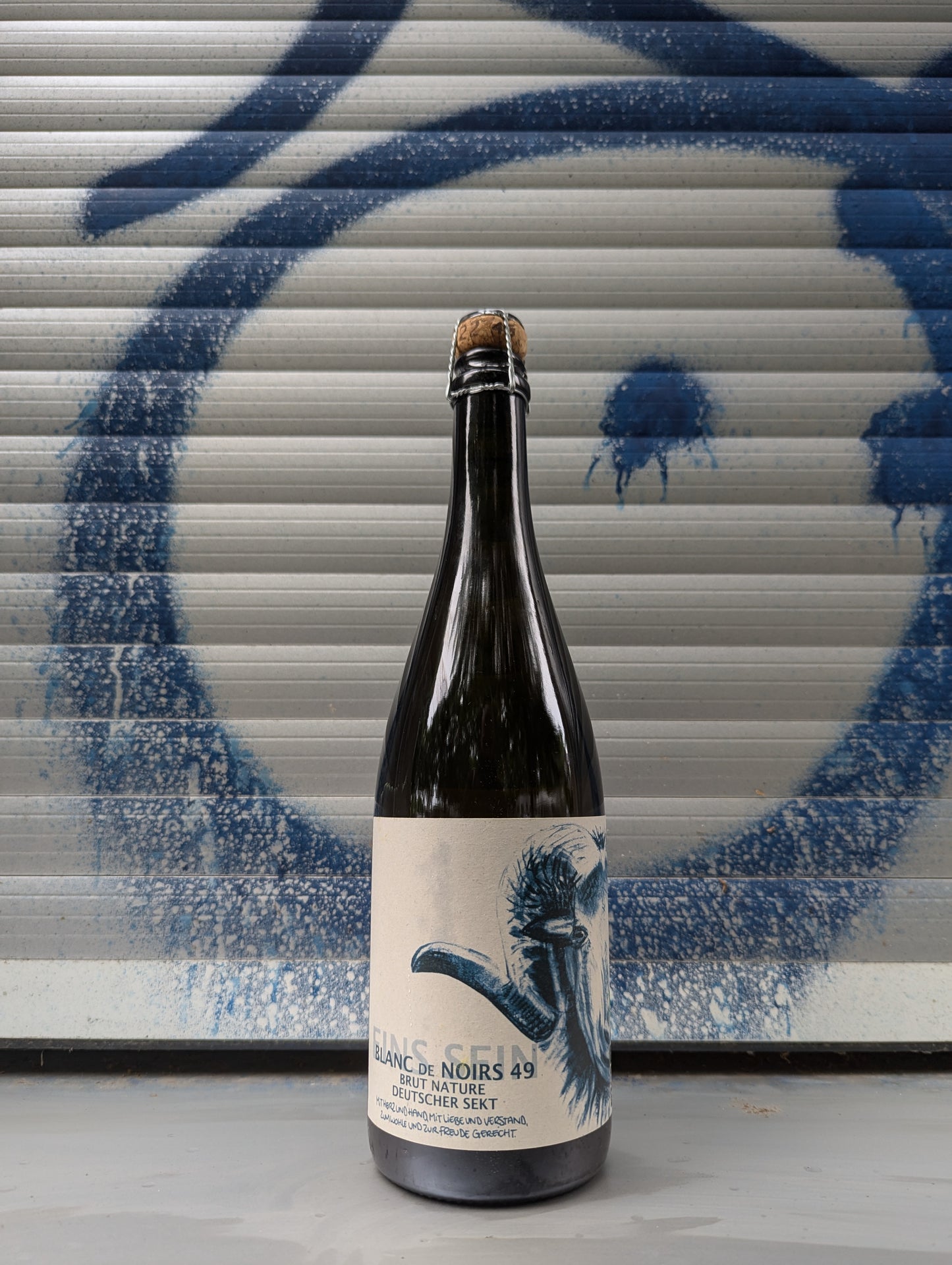 A dark glass bottle sealed with a cork and wire cage. The label reads:
"BLANC DE NOIRS 49"
"BRUT NATURE"
"DEUTSCHER SEKT"
There is a detailed illustration of a ram's head with large, curved horns on the label. The background is a metallic surface with blue graffiti. The text at the bottom of the label appears to be in German and mentions production methods, but it’s partially obscured.