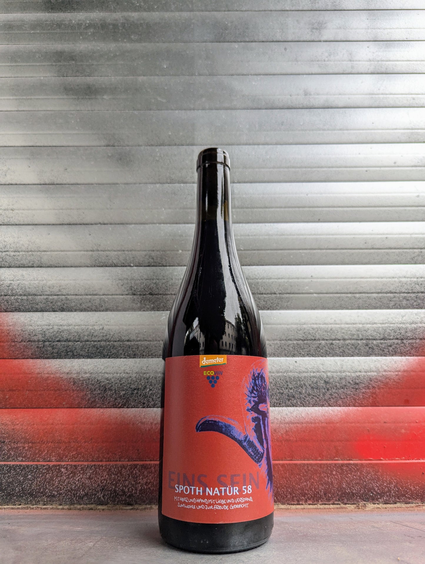 Bottle of Spoth Natur 58 natural wine with a bold red label featuring blue artistic graphics, set against a metallic background with red and black graffiti.