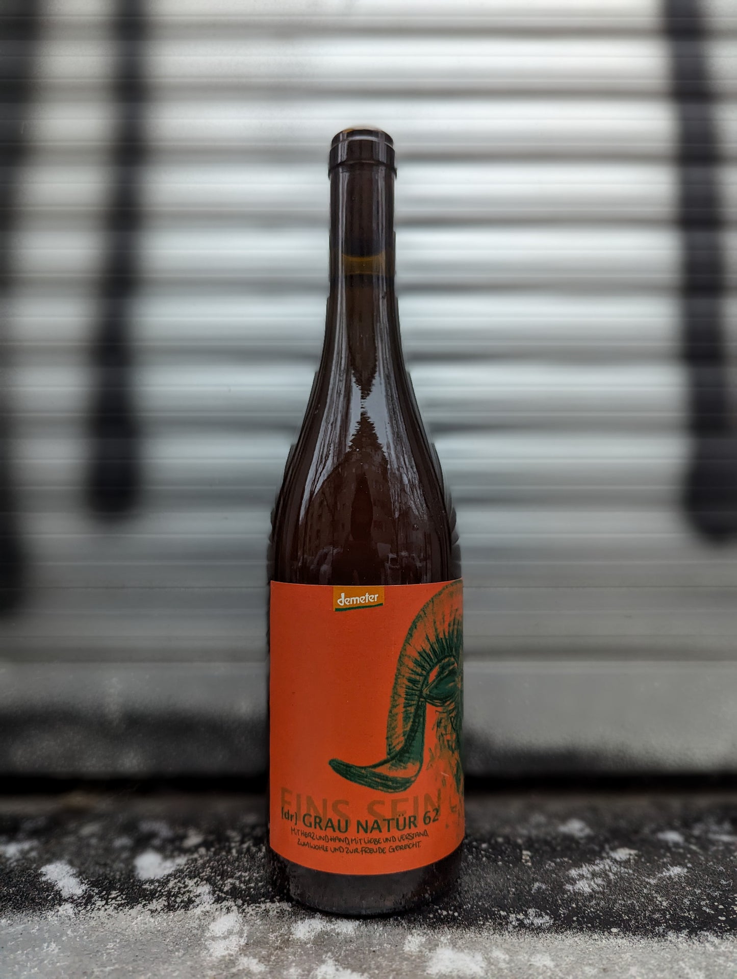 Bottle of Grau Natur 62 natural wine with an orange artistic label, standing against a metallic background with silver and black accents.