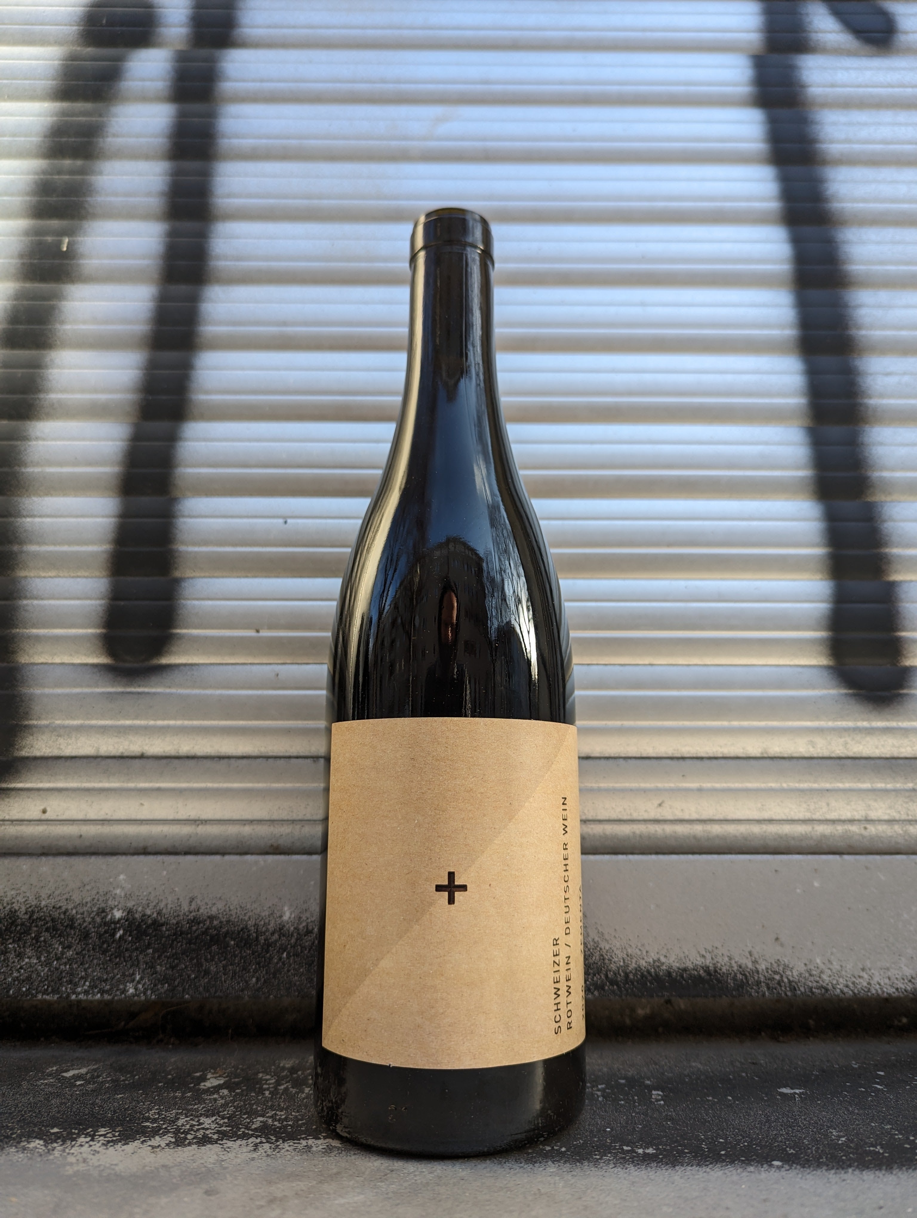 Dark wine bottle with a plain brown label featuring a small black plus sign, set against a metallic, graffiti-marked background.