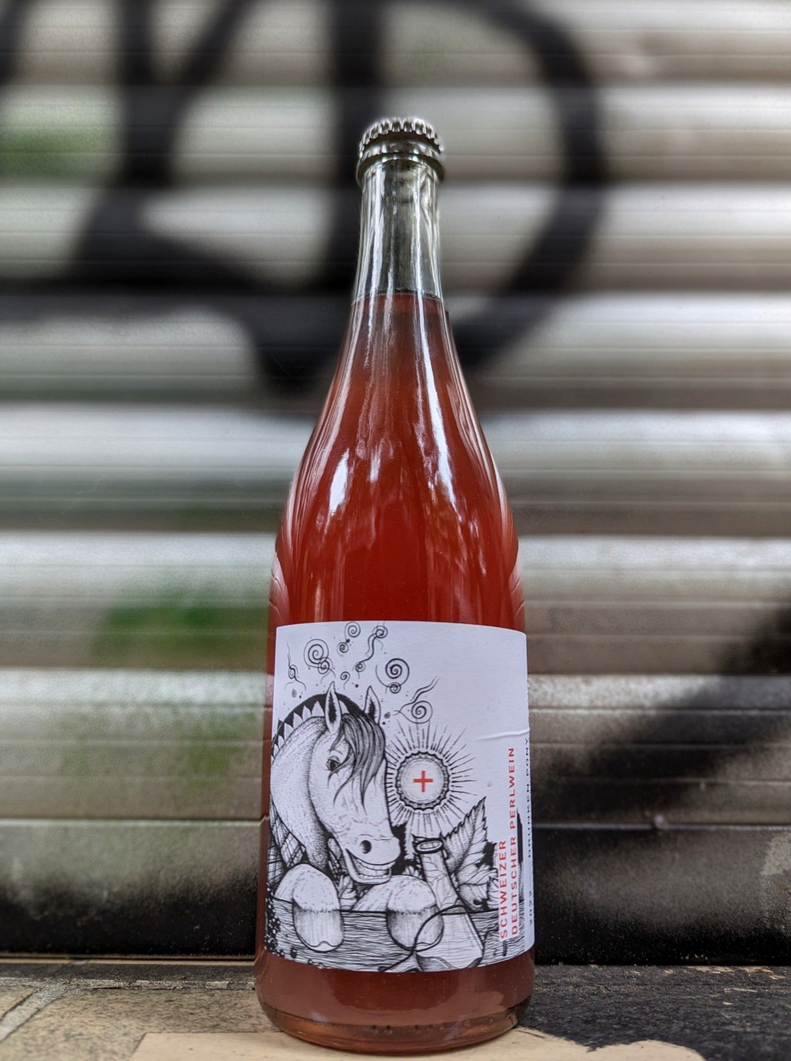 Rosé sparkling wine bottle with an illustrated label featuring a playful horse and sun motif, set against a metallic shutter with graffiti accents in an urban Berlin setting.