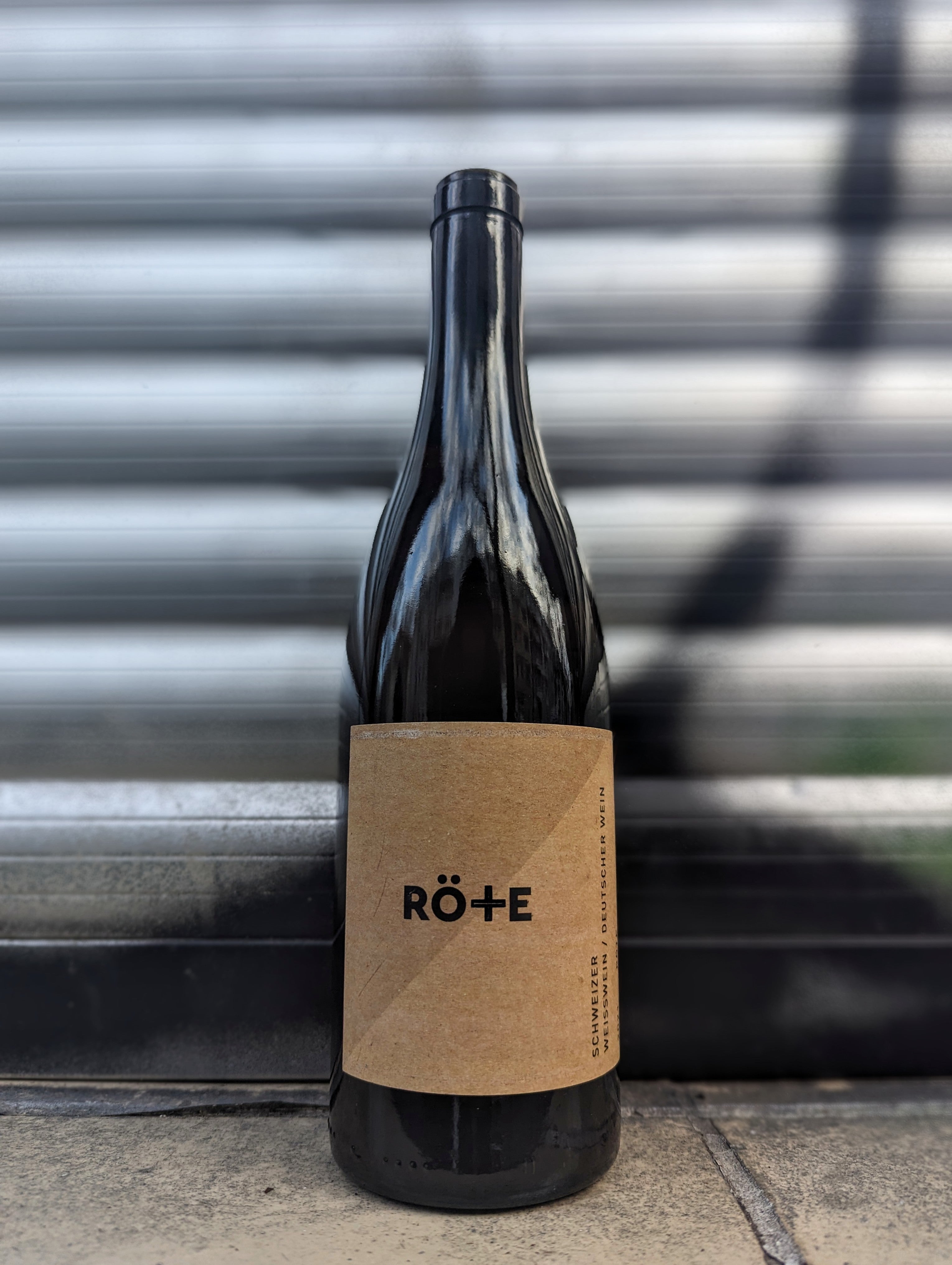 Artisanal natural wine bottle called Röte with a minimalist craft paper label  set against a metallic shutter with graffiti accents in an urban Berlin setting.
