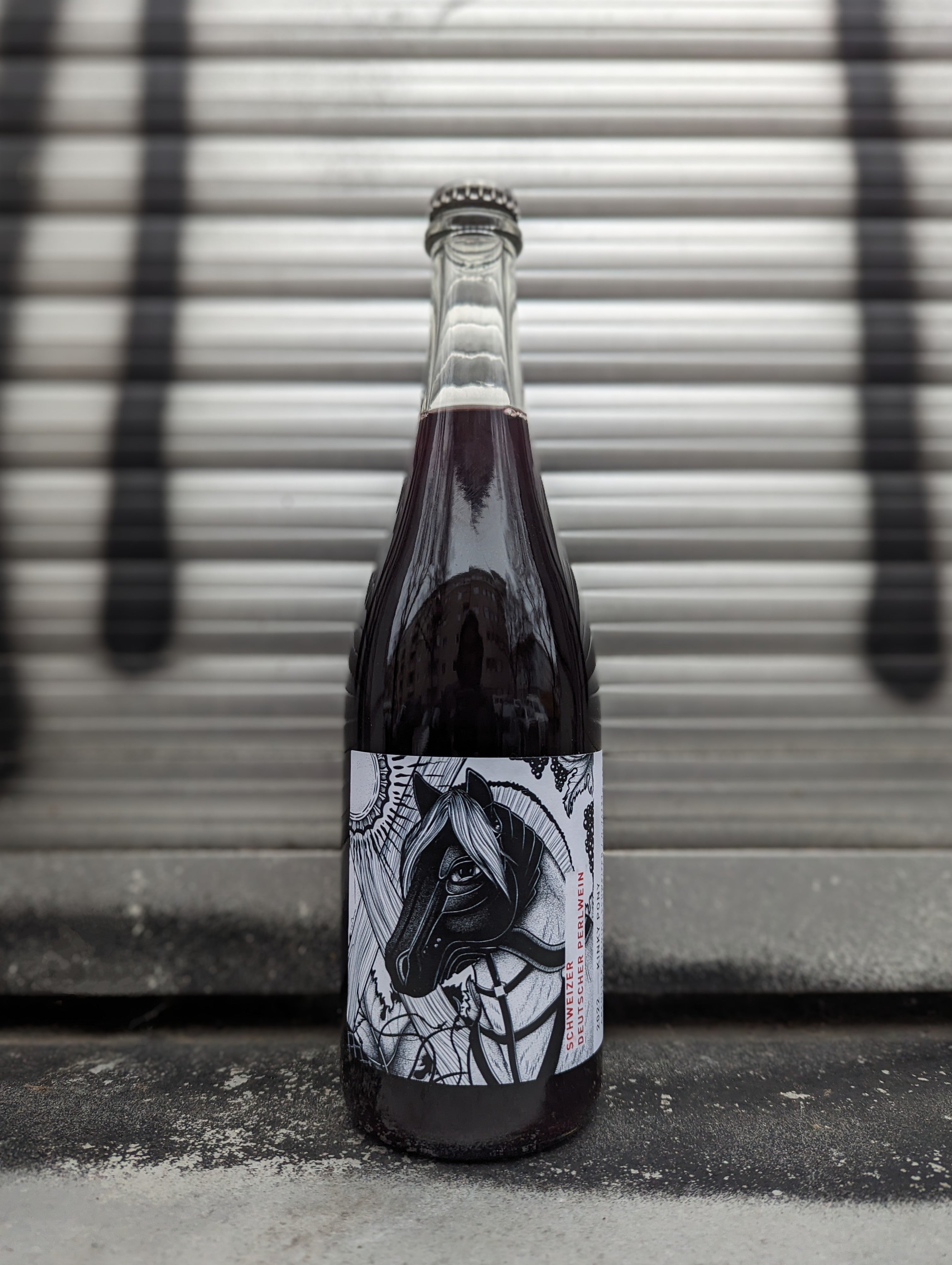 Sparkling red wine bottle with a bold, artistic label featuring a stylized horse illustration in black and white, accented with intricate patterns. Photographed against a metallic shutter backdrop, enhancing the urban aesthetic.