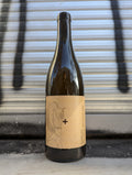 White wine bottle with a minimalist kraft paper label featuring a subtle line drawing of a horse and a bold plus symbol, photographed against an industrial metallic shutter backdrop in an urban setting.