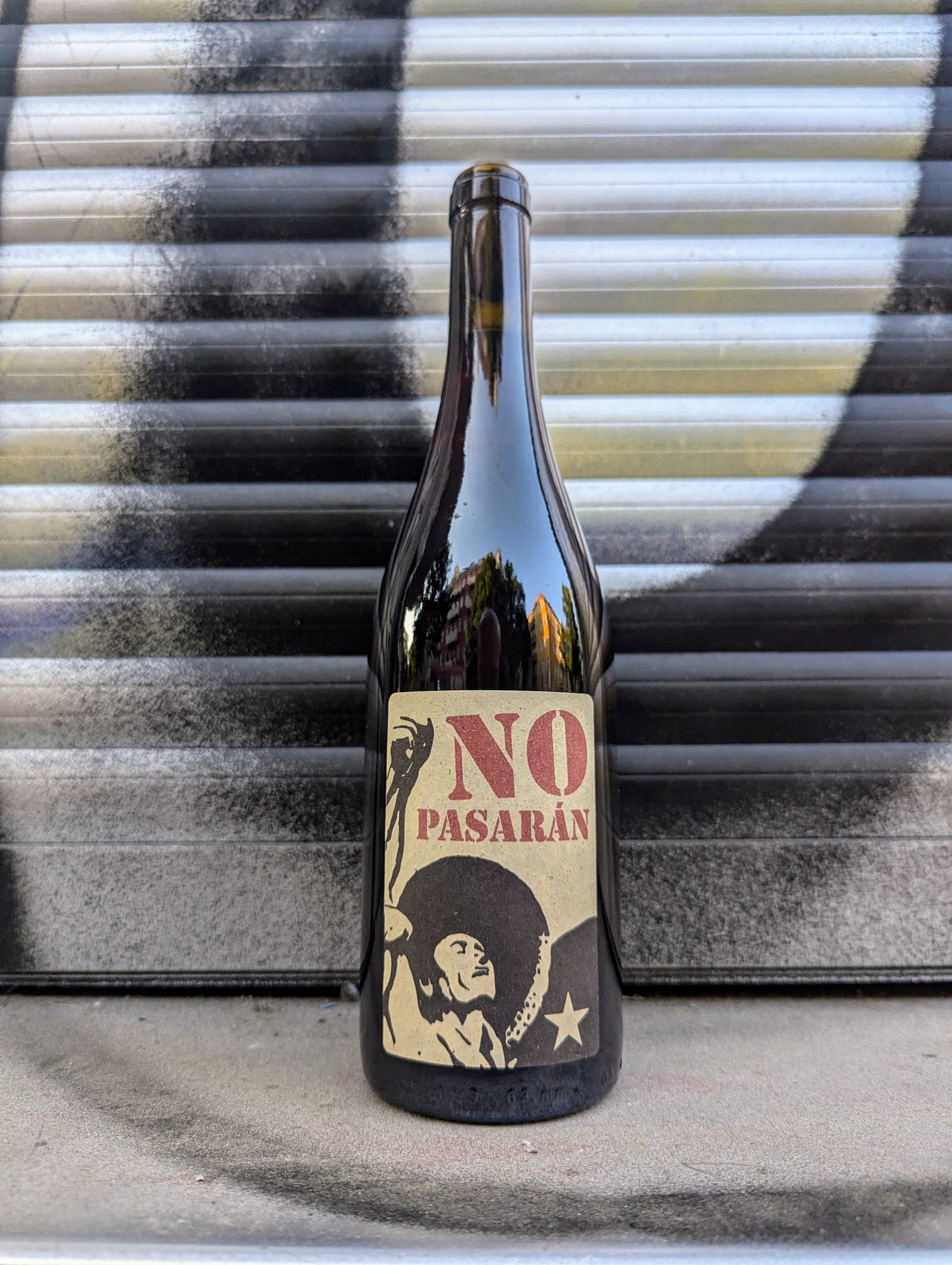 Dark glass wine bottle with a beige label featuring the bold red text "NO PASARÁN" and a black illustration of a person in a hat raising a fist next to a star. The bottle is set against a metallic, industrial background with black and white spray-painted patterns.