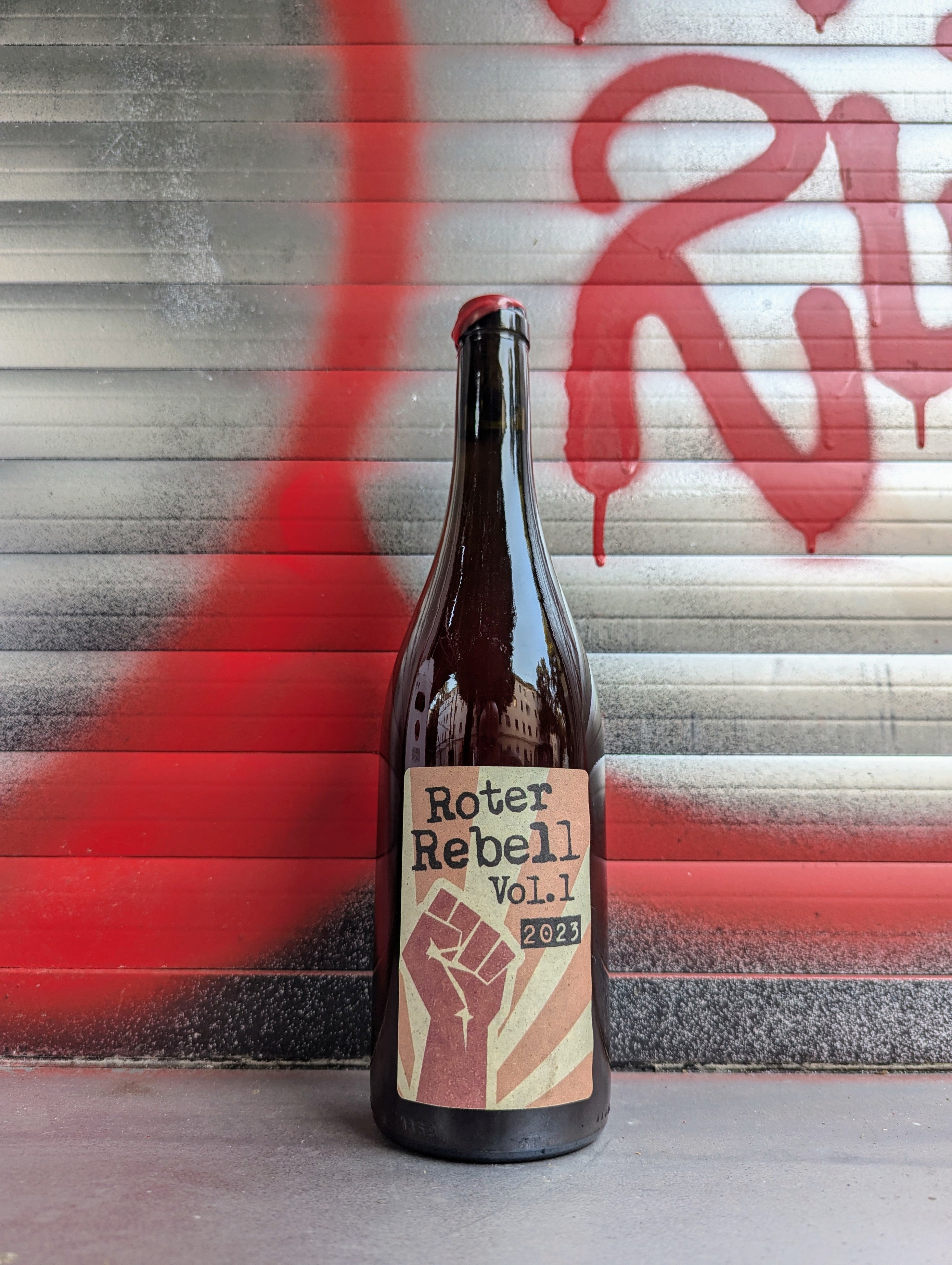 Dark glass wine bottle with a beige and red label displaying the text "Roter Rebell Vol. 1 2023." The label features a bold red raised fist graphic over radiating stripes. The bottle is placed against a metallic background with red and black graffiti, adding an urban and rebellious vibe.