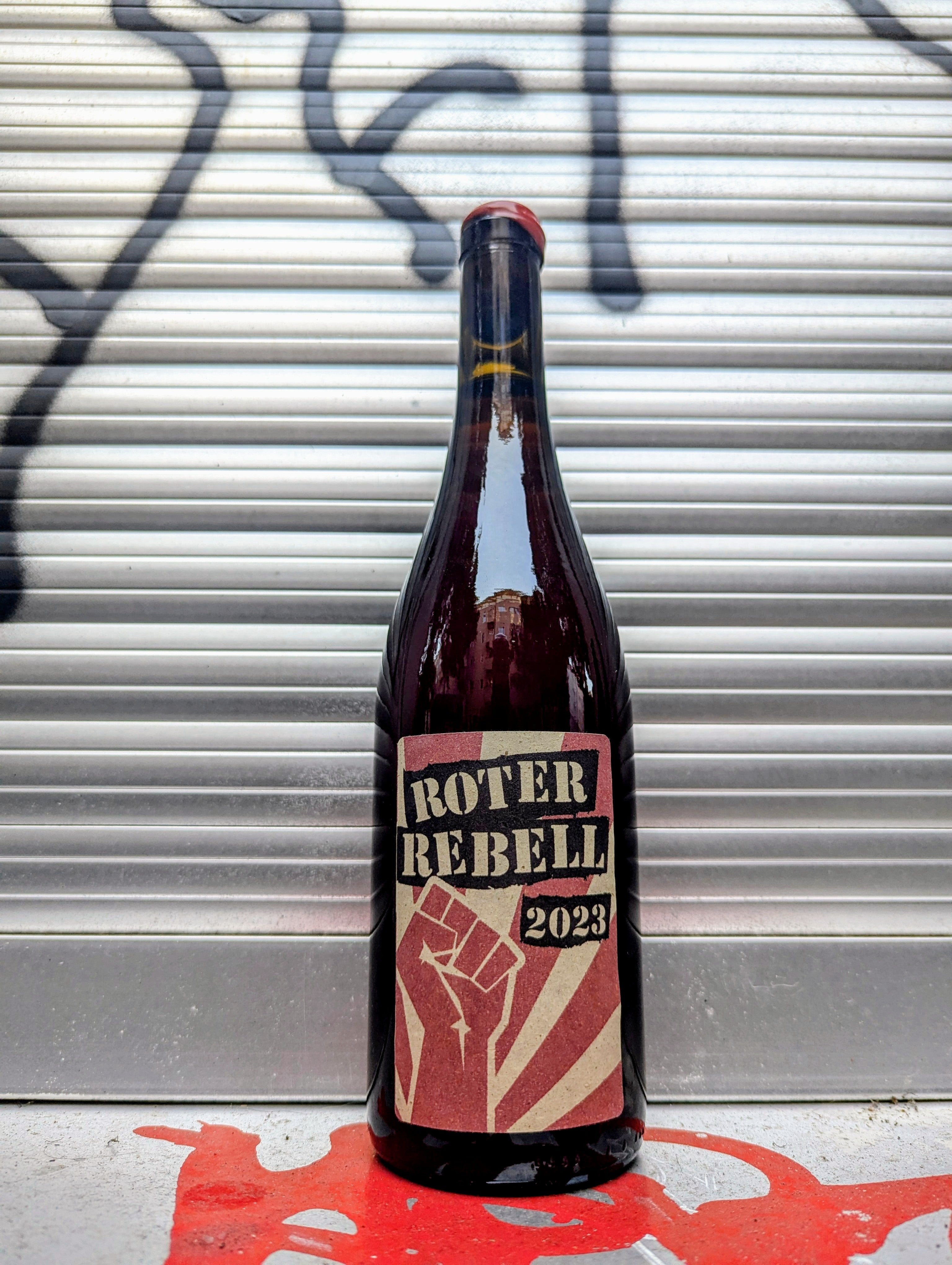 Dark glass wine bottle with a beige and red label displaying the bold black text "ROTER REBELL 2023." The label features a red raised fist graphic over radiating stripes. The bottle is set against a metallic, graffiti-covered background with a red paint splatter on the ground.





