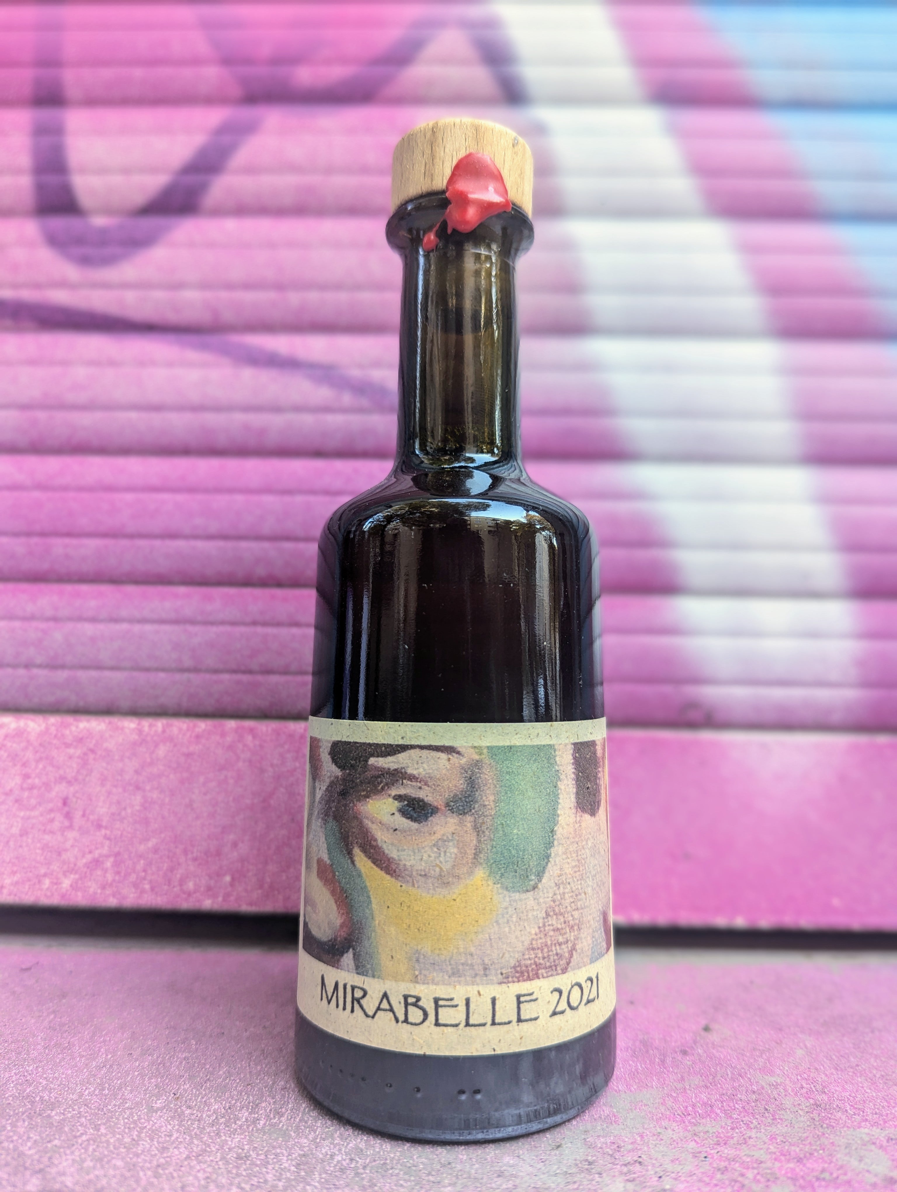 Vinegar bottle called Mirabelle 2021 with a minimalist kraft paper label set against a colorful graffiti-covered shutter in an urban Berlin setting.