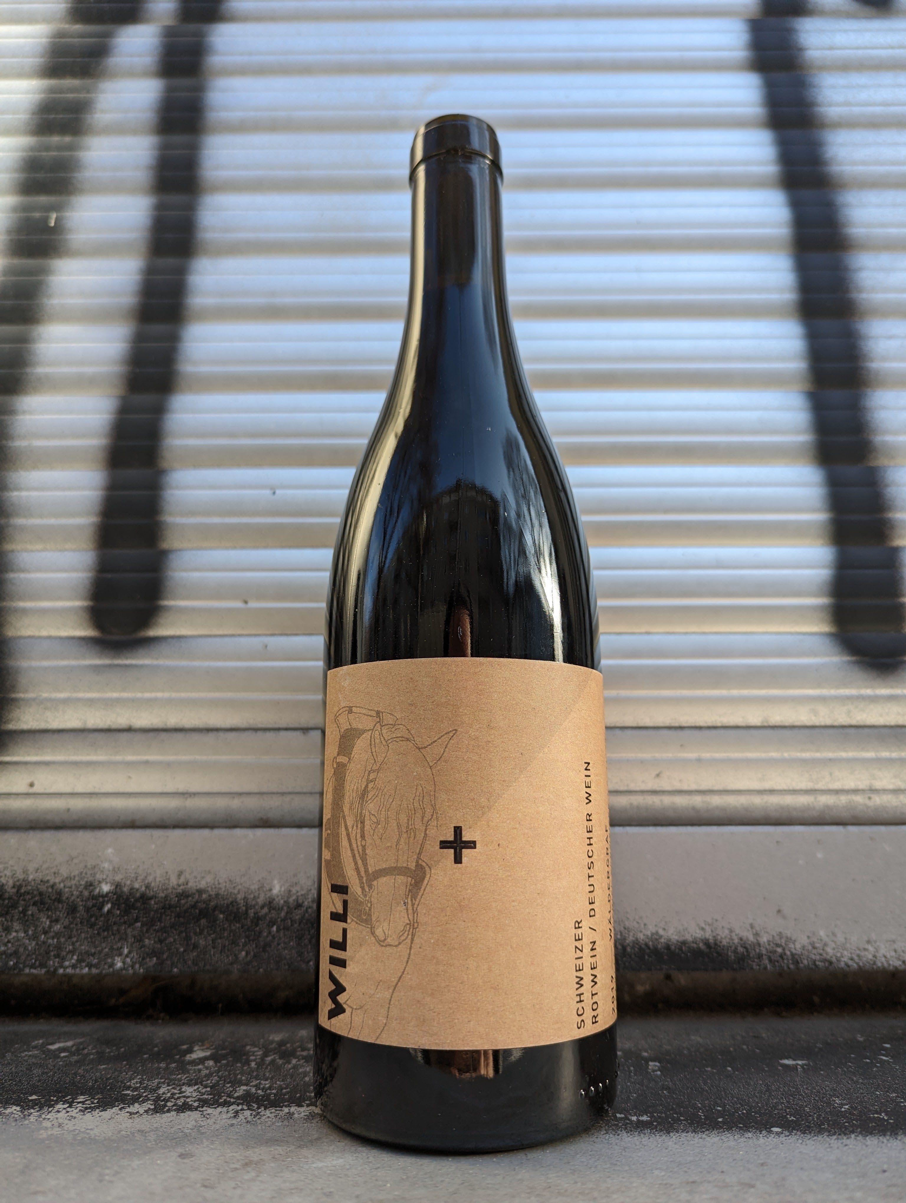 Wine bottle labeled 'Willi' with a minimalist kraft paper design featuring an outline of a horse, placed against a metallic shutter in an urban Berlin environment