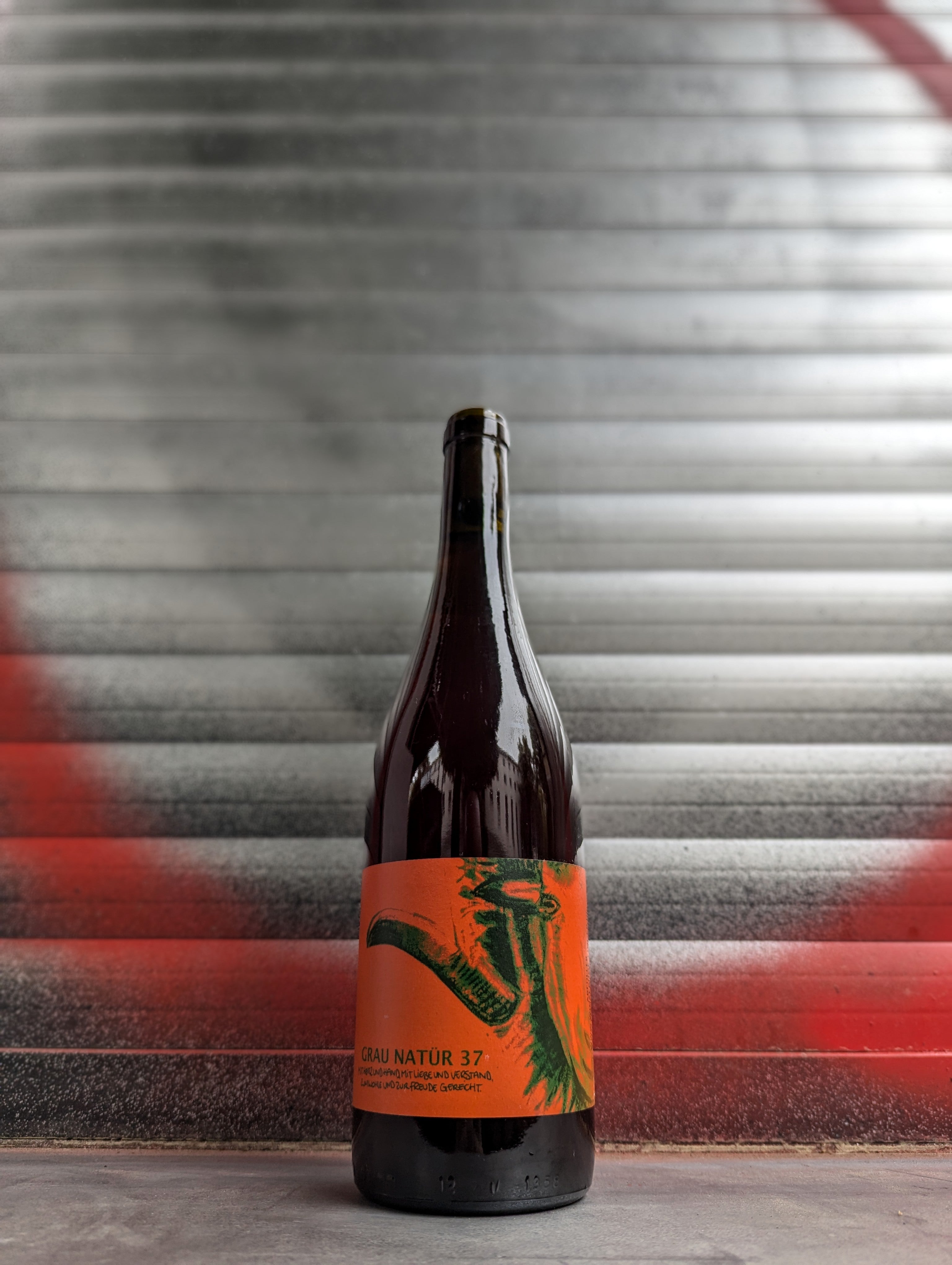 Bottle of Grau Natur 37 natural wine with an orange artistic label, standing against a metallic background with red and black accents.