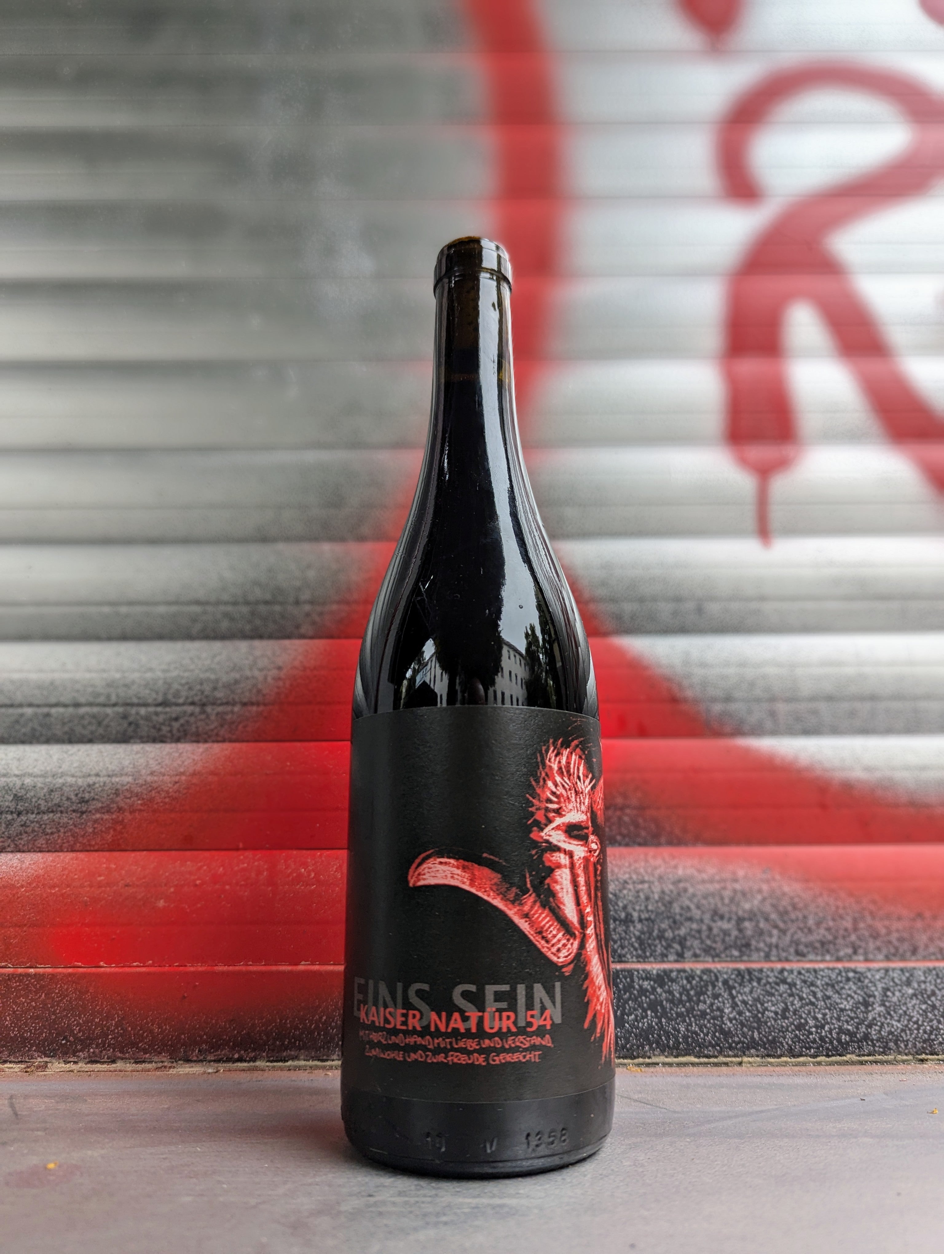 Bottle of Kaiser Natur 54 natural wine with an black and red artistic label, standing against a metallic background with red and silver accents.