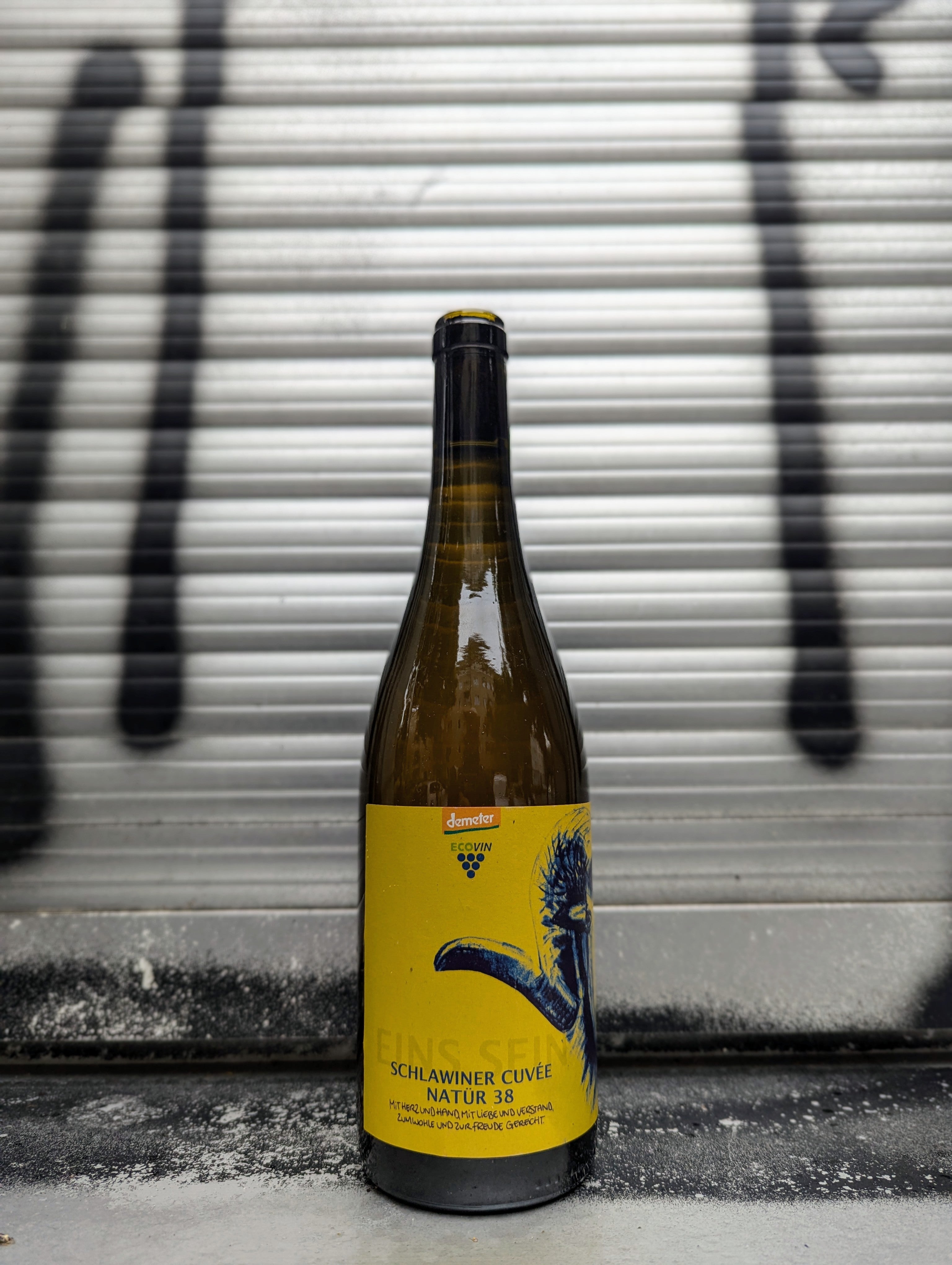 Bottle of Schlawiner Cuvée Natur 38 natural wine with a yellow artistic label, placed against a metallic background with black graffiti accents.