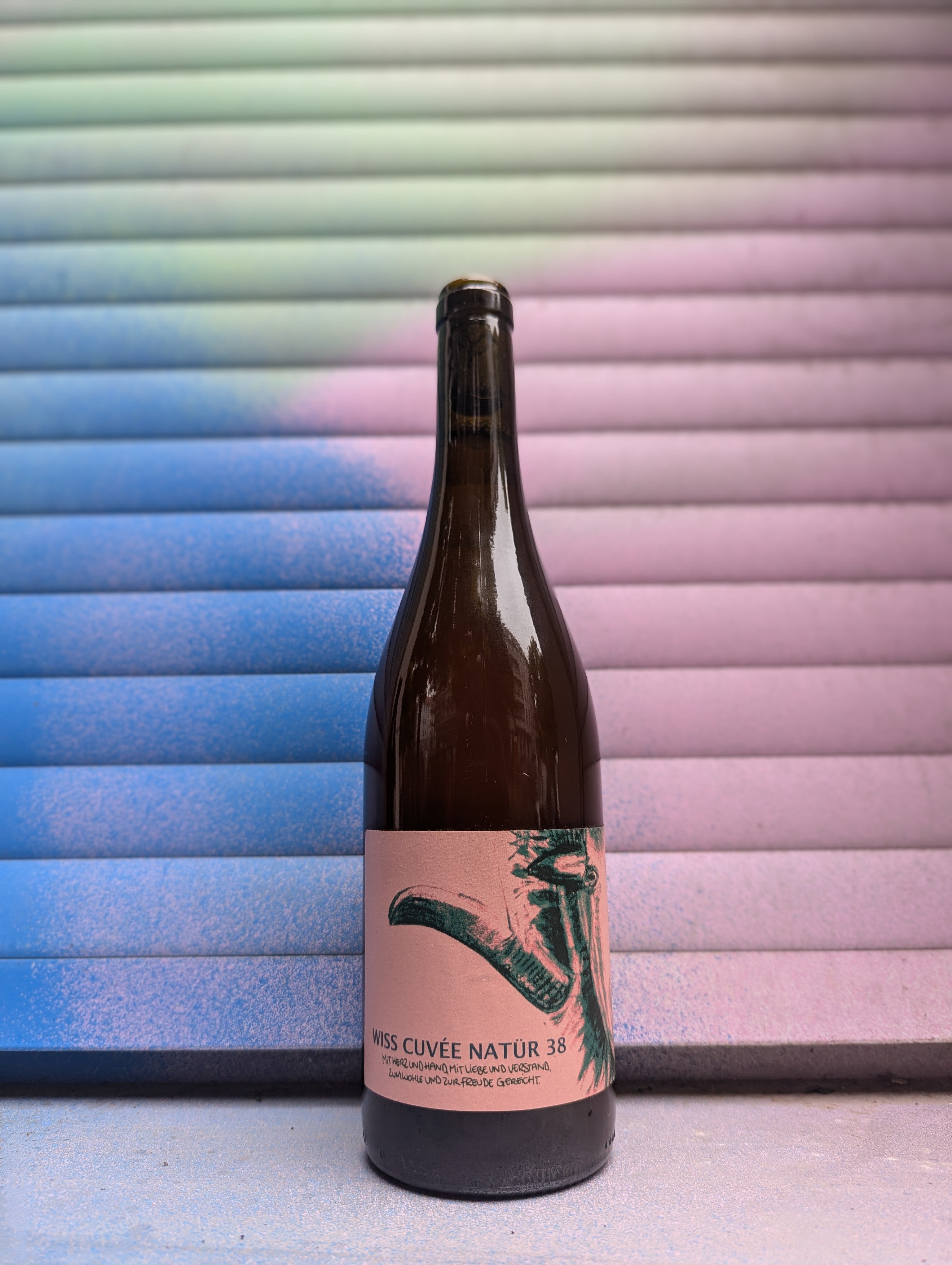 Bottle of Wiss Cuvée Natur 38 natural wine with a soft pink label featuring green artistic graphics, placed in front of a pastel gradient background with shades of blue, pink, and green.